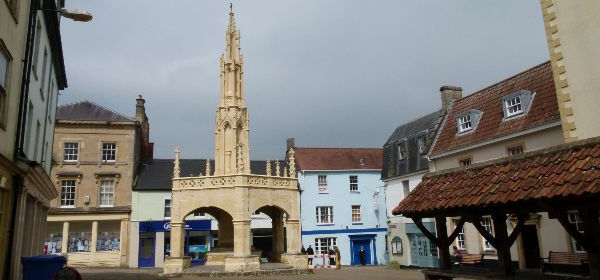 Square surrounded by shows with colourful pastel shaded exterioris. In the centre a warm coloured limestone cross, with shelter underneath.